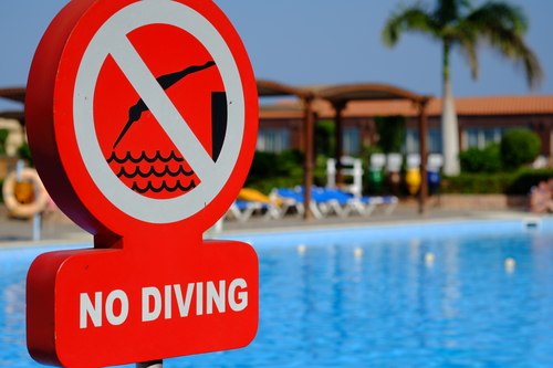Cartel rojo de señalización de prohibido nadar. De fondo, una piscina y reposeras fuera de foco.